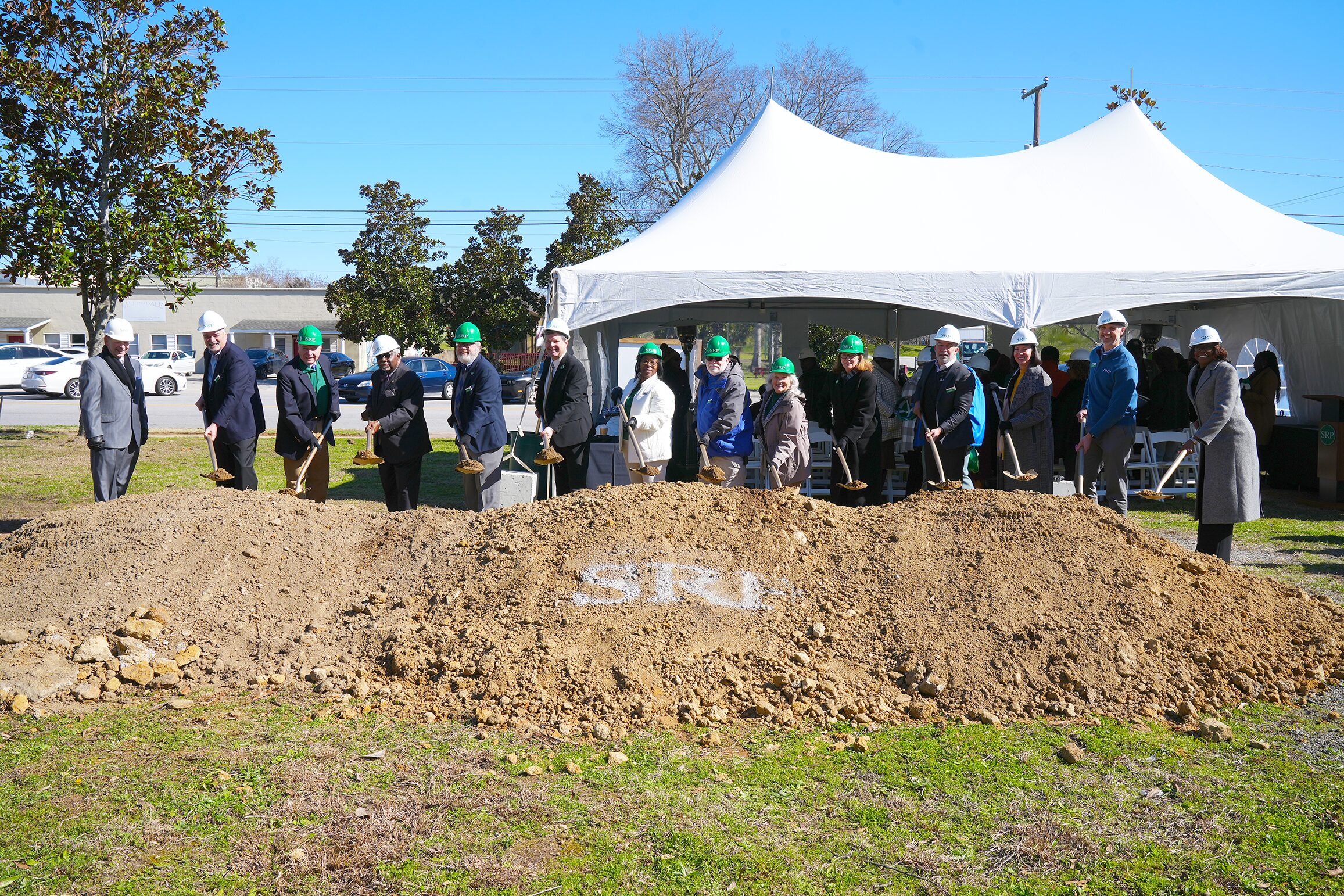 SRP Federal Credit Union board members and executives join local leaders to break ground on the future Allendale branch. The group is lined up behind a dirt mound with the SRP logo painted in the center. The participants are holding shovels.