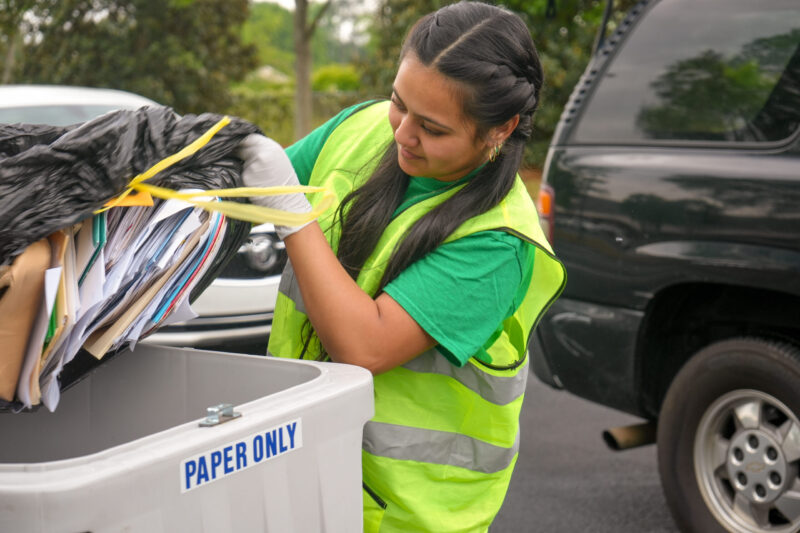 An SRP team member, wearing a safety vest, empties a garbage bag full of paper documents into a bin that reads "Paper Only." Cars in line for Shred Fest are visible in the background.
