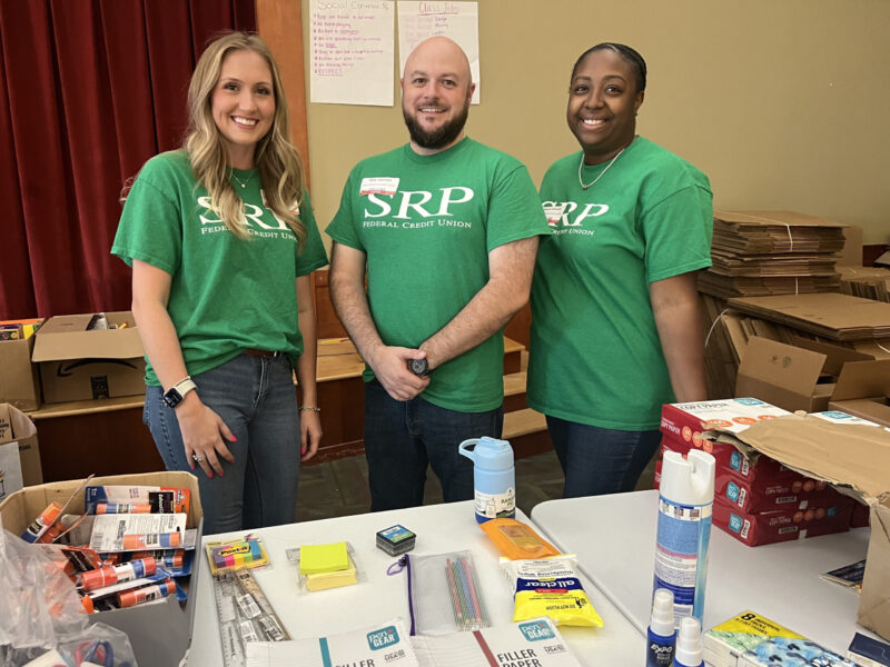 Three SRP employees stand behind a table covered with school supplies.