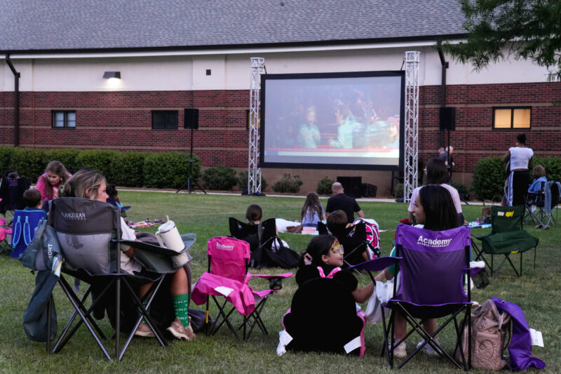 Parents and their children are gathered in front of an outdoor movie screen with lawn chairs and blankets. The movie "Inside Out 2" is on the screen.