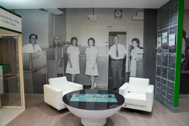 A museum display shows a black-and-white photo of a 1960s SRP branch, a replica of an ATM, logos throughout SRP's history, and an interactive table.