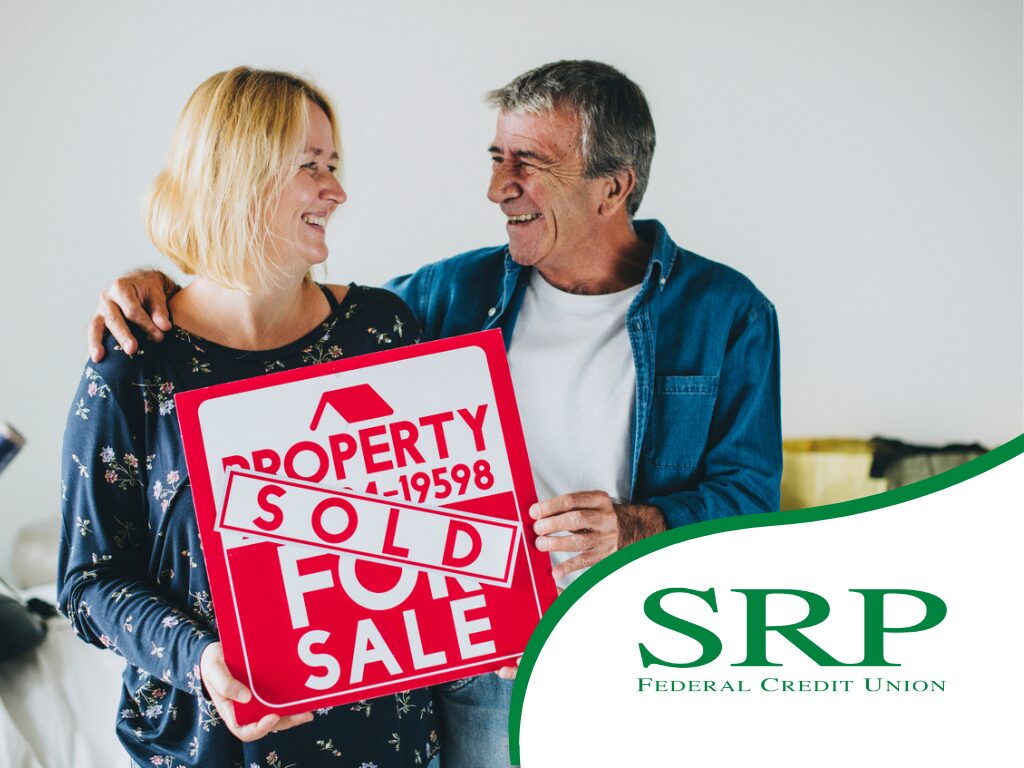 A couple holds a "for sale" sign with a "SOLD" sticker across the surface, representing a couple who decided to downsize their home.