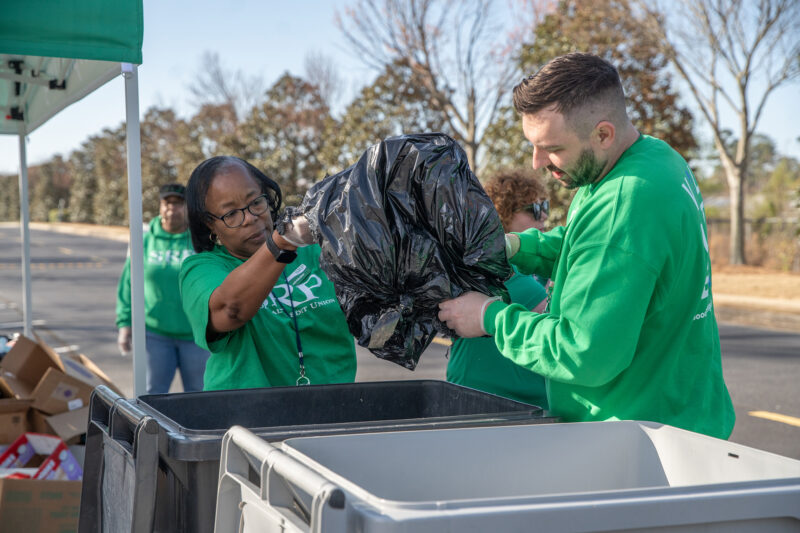 Two people hold a trash bag over a bin.